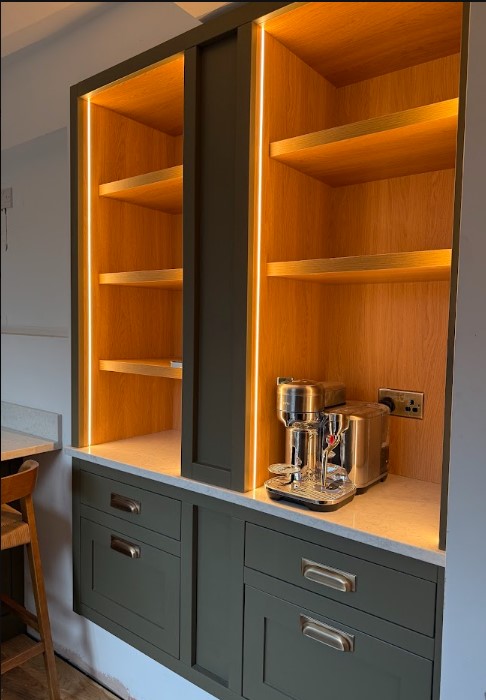 Bespoke in-frame coffee station in deep forest green with warm LED-lit open oak shelving — Hafferty Interiors Cheadle